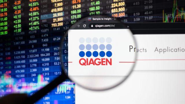 Das Qiagen-Firmenlogo auf einer Website mit verschwommenen Börsenentwicklungen im Hintergrund, gesehen auf einem Computerbildschirm durch ein Vergrößerungsglas