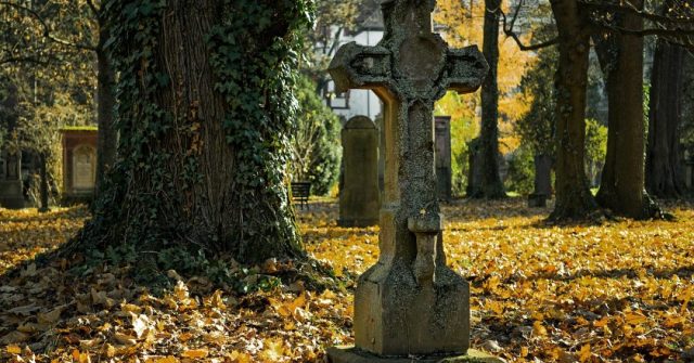 Grabstein in Kreuzform auf einem herbstlichen Friedhof mit großen Baumen und gelben Blättern, die den Boden bedecken.