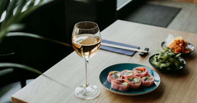 Glas Weißwein, Teller mit Sushi und Essstäbchen auf hellem Holztisch